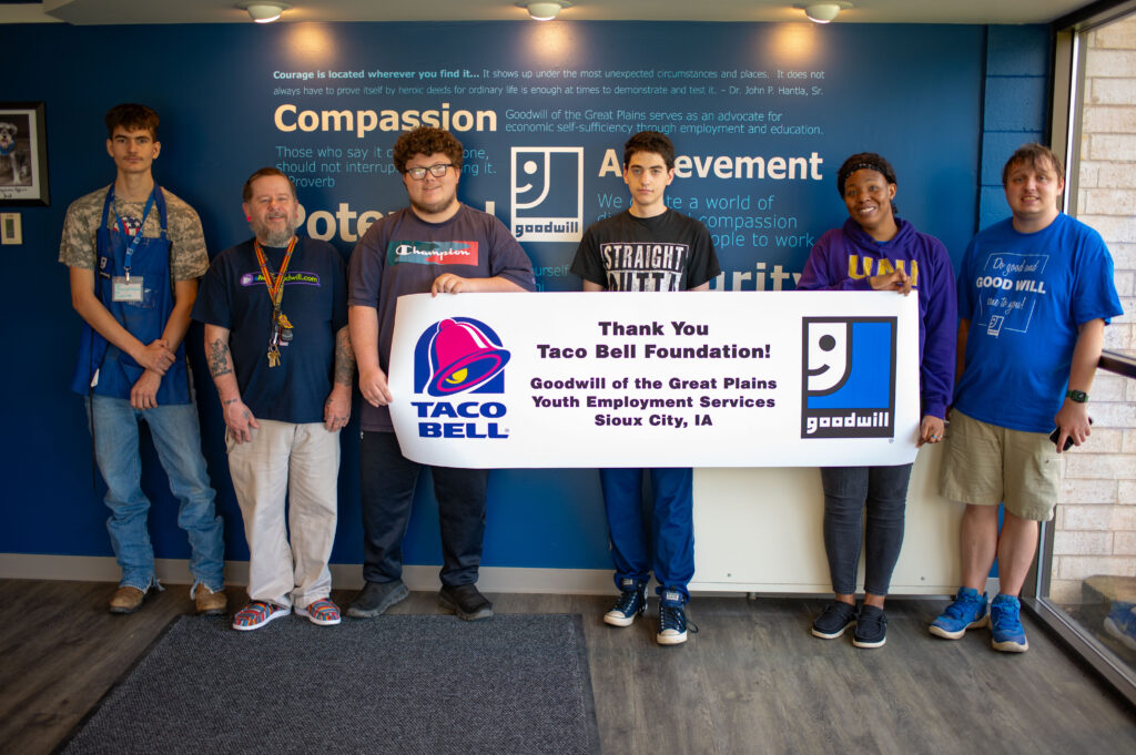Pictured from left to right are Goodwill participants and Job Coach Brendan, Brian, Fred, Charlie, Tatania and Chris, holding a banner which says, "Thank You Taco Bell Foundation! Goodwill of the Great Plains Youth Employment Services, Sioux City, Iowa."

The banner also features a Taco Bell and Goodwill logo.